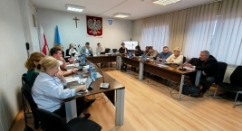 Posiedzenie w sali rady — uczestnicy przy stołach w kształcie litery U, laptopy i mikrofony